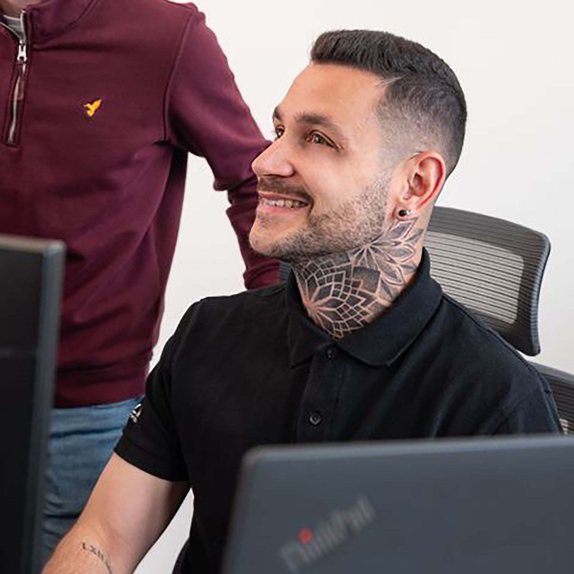 Lächelnder Mann mit Tattoos am Hals, der an einem Computer arbeitet, während eine Person im Hintergrund steht.