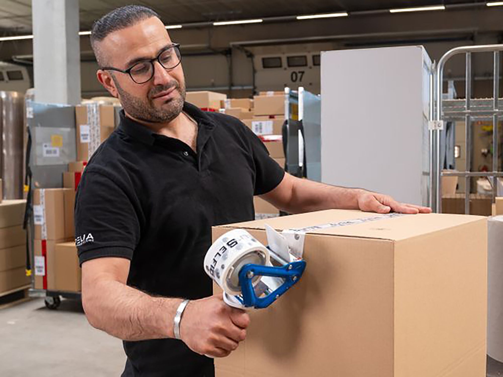 Mann mit Brille, der ein Paket mit einem Klebebandabroller verschließt, in einem Lager.