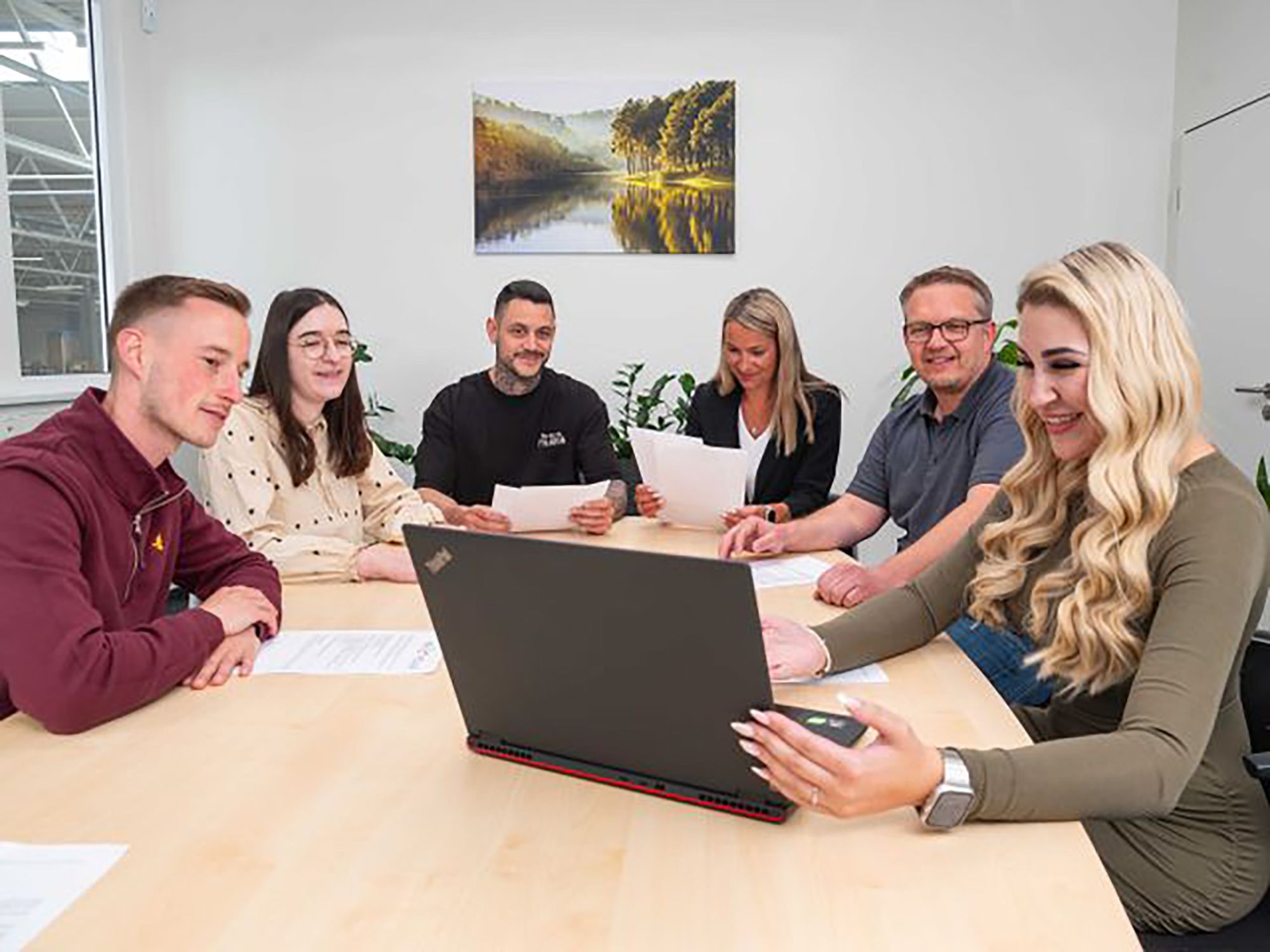 Sechs Personen sitzen an einem Tisch, schauen auf einen Laptop und halten Papiere in der Hand. Im Hintergrund ist ein Bild mit einer Landschaft zu sehen.