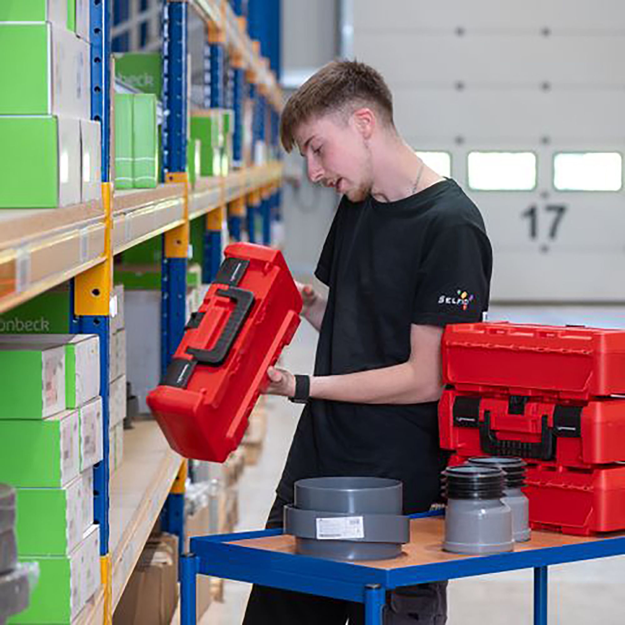 Junger Mann in einem Lager, der eine rote Werkzeugkiste von einem Regal nimmt.