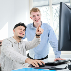 Zwei junge Männer arbeiten gemeinsam am Computer in einem hellen Büro. Einer sitzt vor dem Bildschirm und bedient die Maus, während der andere daneben steht, lächelt und auf den Monitor zeigt, als würde er etwas erklären oder hervorheben.