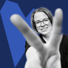 Eine lächelnde Frau mit Brille zeigt ein Peace-Zeichen in die Kamera; das Bild ist in Schwarz-Weiß gehalten mit blauem grafischen Hintergrund.