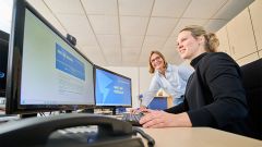 Zwei Frauen arbeiten gemeinsam an einem Schreibtisch im Büro, schauen auf zwei Monitore und besprechen Inhalte, während eine die Maus bedient und die andere daneben steht.