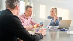 Drei Personen sitzen in einem hellen Büro an einem Tisch, besprechen Unterlagen und arbeiten mit Laptops, während sie sich konzentriert austauschen.