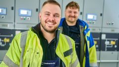 Zwei Mitarbeitende in Warnschutzkleidung stehen in einer technischen Anlage vor Schalttafeln; die Person im Vordergrund lächelt in die Kamera, während die zweite im Hintergrund steht und ebenfalls lächelt.