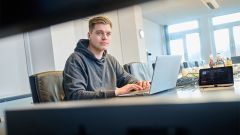 Ein junger Mann sitzt in einem modernen Büro an einem Tisch und arbeitet konzentriert an einem Laptop; daneben liegen Kabel und technische Geräte.