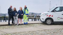 Vier Mitarbeitende stehen gemeinsam an einem Hafen und schauen in die Kamera; neben ihnen ist ein Firmenfahrzeug zu sehen, im Hintergrund Boote und Stege.