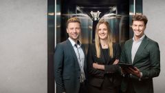 Drei junge Business-Profis in eleganter Kleidung stehen gemeinsam lächelnd vor einem modernen Aufzug.