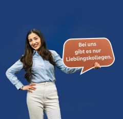 Eine lächelnde Frau mit langen dunklen Haaren, trägt eine blaue Bluse und eine helle Hose. Sie hält eine Sprechblase mit der Aufschrift "Bei uns gibt es nur Lieblingskollegen" in der Hand.