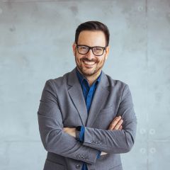 Ein lächelnder Mann mit Brille und Bart, trägt ein graues Sakko über einem blauen Hemd. Er steht mit verschränkten Armen vor einer hellgrauen Wand und wirkt freundlich und selbstbewusst.