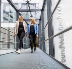 Zwei lächelnde Frauen gehen Seite an Seite durch einen modernen, hellen Glasgang in einem Bürogebäude. Die Frau links trägt eine gestreifte Jacke über einem schwarzen Oberteil und schwarze Jeans. Die Frau rechts trägt eine blaue Jacke über einem schwarzen Oberteil und schwarze Jeans. An der rechten Wand ist ein weißer Schriftzug zu erkennen, der den Begriff "VerbundVolksbank OWL" enthält. Das Bild vermittelt eine positive und professionelle Arbeitsatmosphäre.