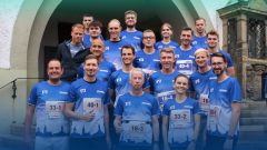 Eine große Gruppe lächelnder Personen, etwa 20 Männer und 3 Frauen, steht eng zusammen und blickt in die Kamera.  Alle tragen ein blaues Sport-T-Shirts von der VerbundVolksbank OWL  sowie Startnummern für einen Laufwettbewerb. Am unteren Bildrand steht in weißer Schrift der Text "Werde Teil des Teams." Das Bild symbolisiert Teamgeist, Zusammenhalt und Engagement, im Rahmen eines Firmenlaufs.