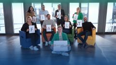 Eine Gruppe von neun lächelnden Personen steht und sitzt in einem modernen Büroumfeld und hält einzelne weiße Schilder hoch, die zusammen das Wort "VIELFALT" buchstabieren. In der vorderen Reihe sitzt eine Frau in grüner Jacke auf dem Boden und hält ein größeres weißes Schild mit dem Logo "Charta der Vielfalt – Unterzeichnet" hoch. Im Hintergrund sind große Fenster und ein helles, offenes Büro zu sehen. Am unteren Bildrand steht in weißer Schrift der Text "Werde Teil des Teams." Das Bild symbolisiert die Wertschätzung von Vielfalt und Inklusion innerhalb des Unternehmens.