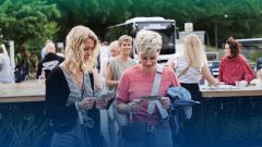Zwei Frauen stehen im Vordergrund und blicken konzentriert auf zwei Eintrittskarten, die sie in den Händen halten. Die Frau links hat lange, blonde Locken und trägt ein gestreiftes Oberteil mit V-Ausschnitt. Die Frau rechts hat kurze, blonde Haare und trägt ein rosa T-Shirt mit einem gemusterten Trägerband über der Schulter. Im unscharfen Hintergrund sind weitere Personen und ein Bus zu sehen, was auf eine Veranstaltung im Freien hindeutet. Am unteren Bildrand steht in weißer Schrift der Text "Werde Teil des Teams." Das Bild vermittelt eine entspannte und interaktive Atmosphäre, die den Teamzusammenhalt fördert.