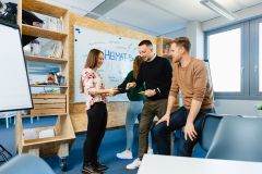 Gruppe junger Menschen arbeitet vor Whiteboard mit dem Begriff „HEIMAT-Banker“ in kreativem Büro.