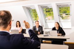 Vier Personen in Besprechung mit Laptops, eine Hand gestikuliert im Vordergrund, Dachfenster im Hintergrund.