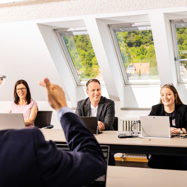 Vier Personen in Besprechung mit Laptops, eine Hand gestikuliert im Vordergrund, Dachfenster im Hintergrund.