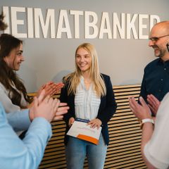 Junge Frau wird vor „HEIMATBANKER“-Schriftzug für ein Zertifikat von Kolleg:innen beklatscht.