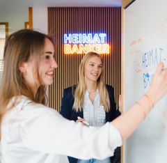 Zwei junge Frauen schreiben an ein Whiteboard, im Hintergrund leuchtet „HEIMAT BANKER“.