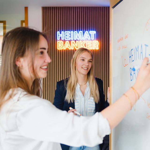 Zwei junge Frauen schreiben an ein Whiteboard, im Hintergrund leuchtet „HEIMAT BANKER“.