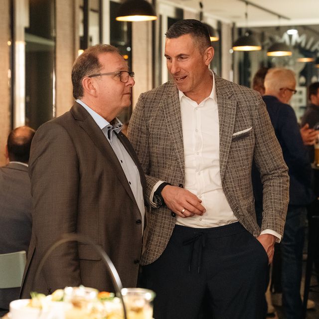 Zwei Männer im Anzug unterhalten sich bei einer Abendveranstaltung mit Buffet im Hintergrund.