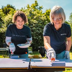 Zwei Frauen streichen Holzbretter im Freien, tragen Handschuhe und VR Bank-Shirts.