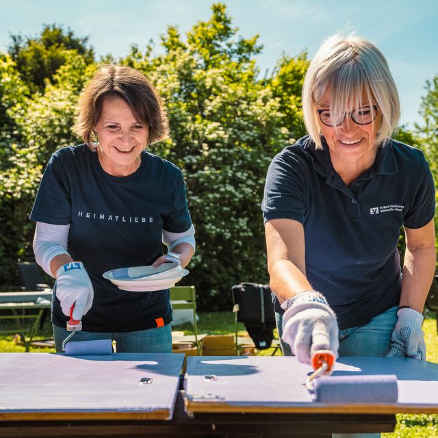Zwei Frauen streichen Holzbretter im Freien, tragen Handschuhe und VR Bank-Shirts.