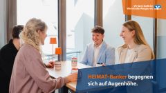 Vier Personen lachen im Gespräch an einem Tisch mit HEIMAT-Banker-Tassen, Text: „HEIMAT-Banker begegnen sich auf Augenhöhe“.