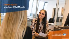 Beraterin am Schalter lächelt Kundin an, Text: „HEIMAT-Banker schreiben SERVICE groß.