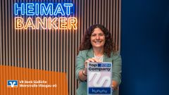 Frau hält Kununu-Siegel „Top Company 2024“ vor „HEIMAT-BANKER“-Leuchtschrift der VR Bank Südliche Weinstraße-Wasgau.